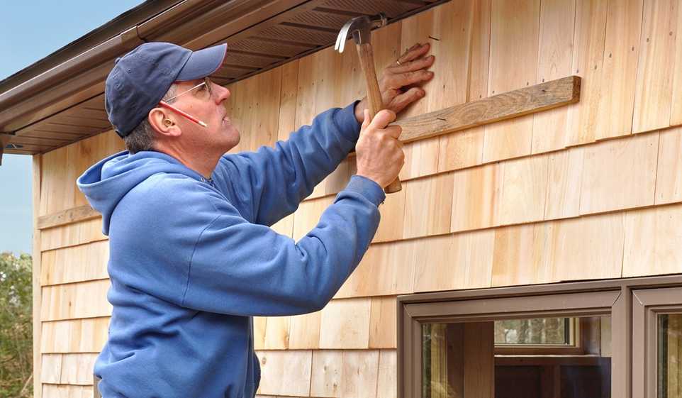 Contractor wearing blue sweatshirt, using hammer to nail a nail into a wooden shingle