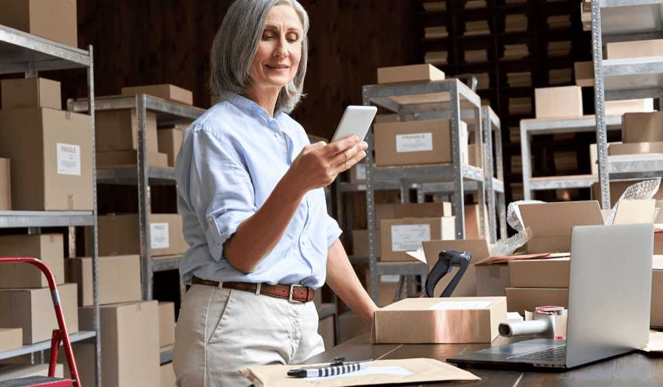 A standing in a storage room of boxes looks at a smartphone. She stands in front of a laptop