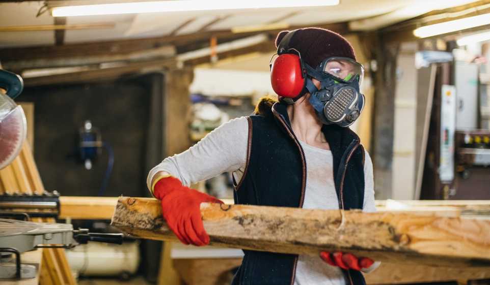 Woman wearing goggles and gas mask carrying wooden beam.