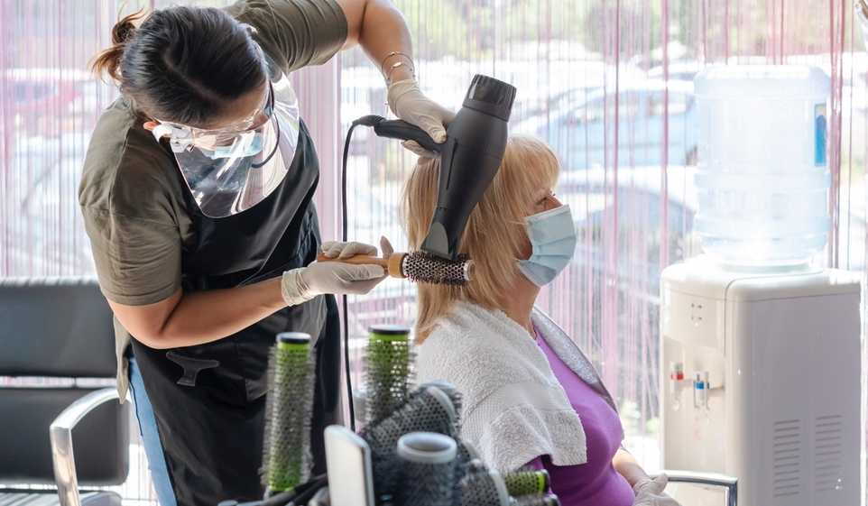 One woman, a hairstylist, blow drying another woman's hair. Both women are wearing face masks.