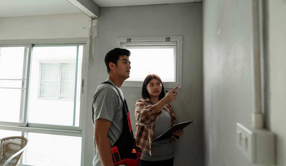 Man and woman looking at a wall during a project. Woman pointing to wall, man wearing tool belt.