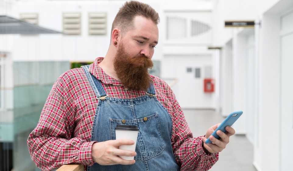 Man wearing button down shirt under blue overalls, carrying a coffee in one hand and smartphone in the other.