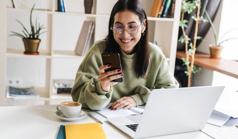 Woman wearing glasses holding iPhone in front of laptop while drinking a cup of coffee.