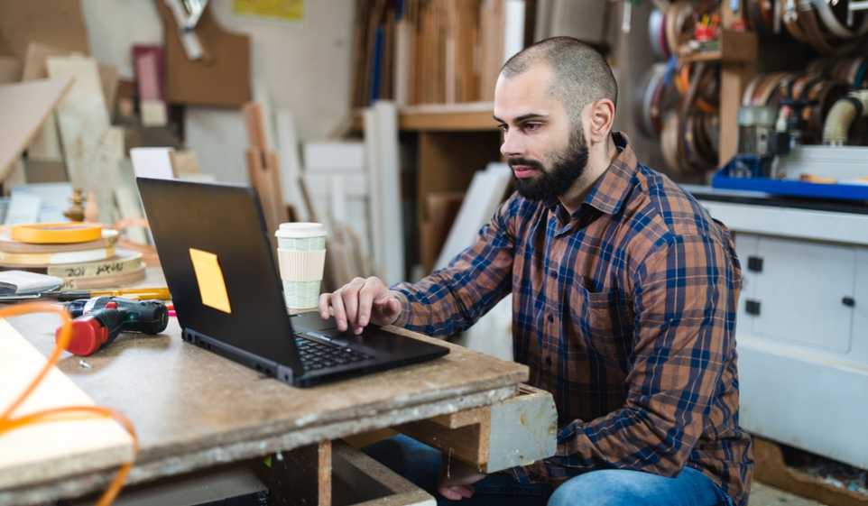 Man wearing plaid shirt, sitting in workshop while working on a black laptop.