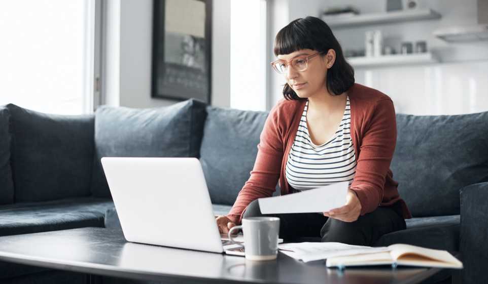 Woman with glasses, sitting on couch looking at paper and laptop.