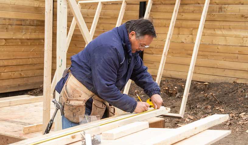 Michigan builder in navy sweatshirt measuring a piece of wood with a tape measure and marking it with pencil