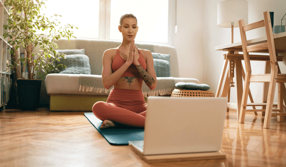Woman is yoga outfit sits on mat in front of an open laptop.