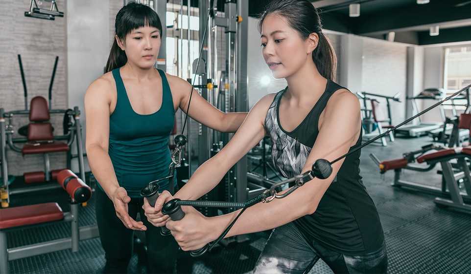 A personal trainer helps her female client with resistance training in a gym