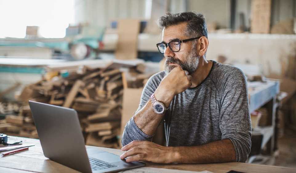 A bearded man wearing glasses ponders over his laptop.