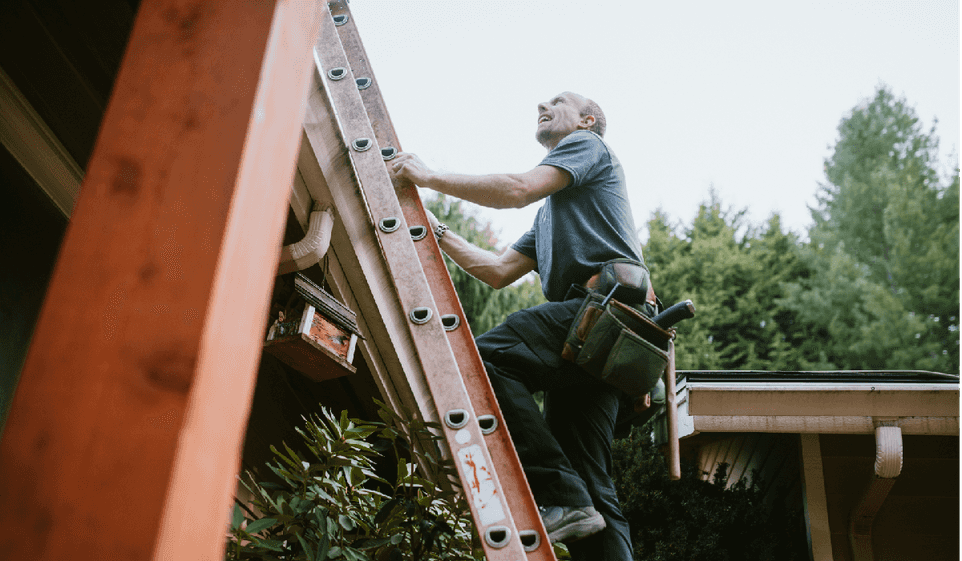 A man in a t-shirt climbs a ladder on the side of a house.