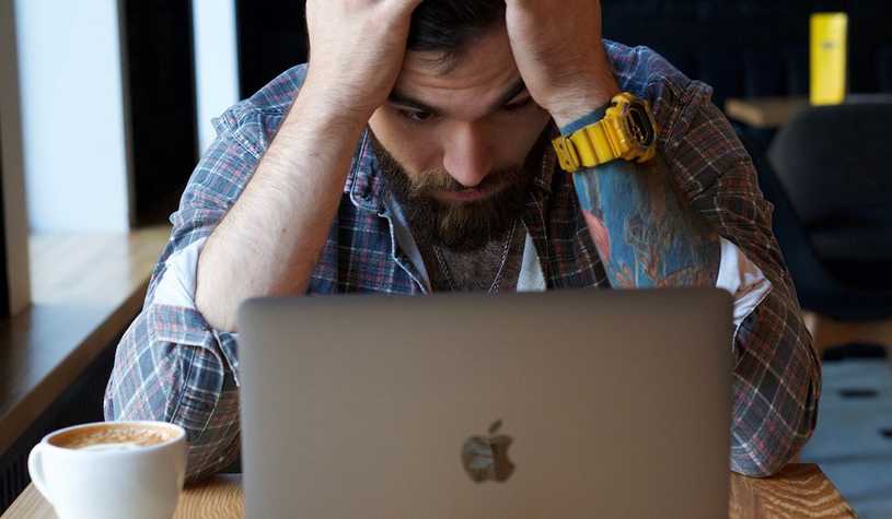 Man sitting at desk in front of a laptop with his dead in his hands, looking distraught.