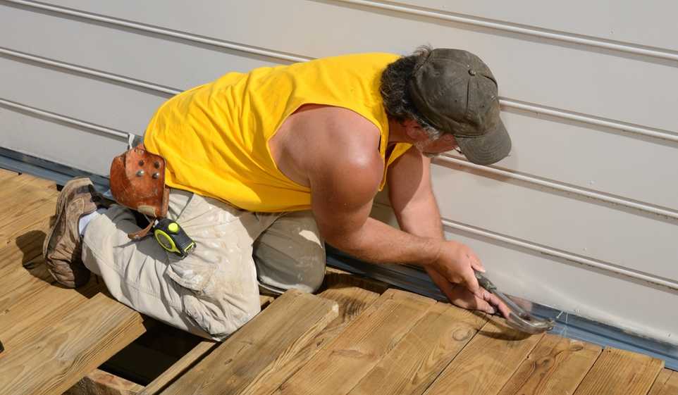 Carpenter wearing yellow shirt, kneeling on wooden deck while hammering into the side of a house.