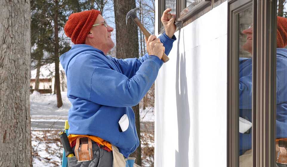 Man in blue sweatshirt and red hat, wearing glasses, using hammer on the side of the house