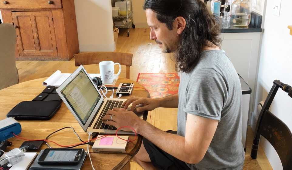 Man with long hair, sitting at table in his home, working on a laptop.