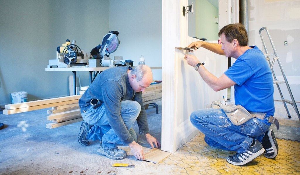 Contractor kneeling, using scissors to cut tape to measure border before painting, working alongside coworker