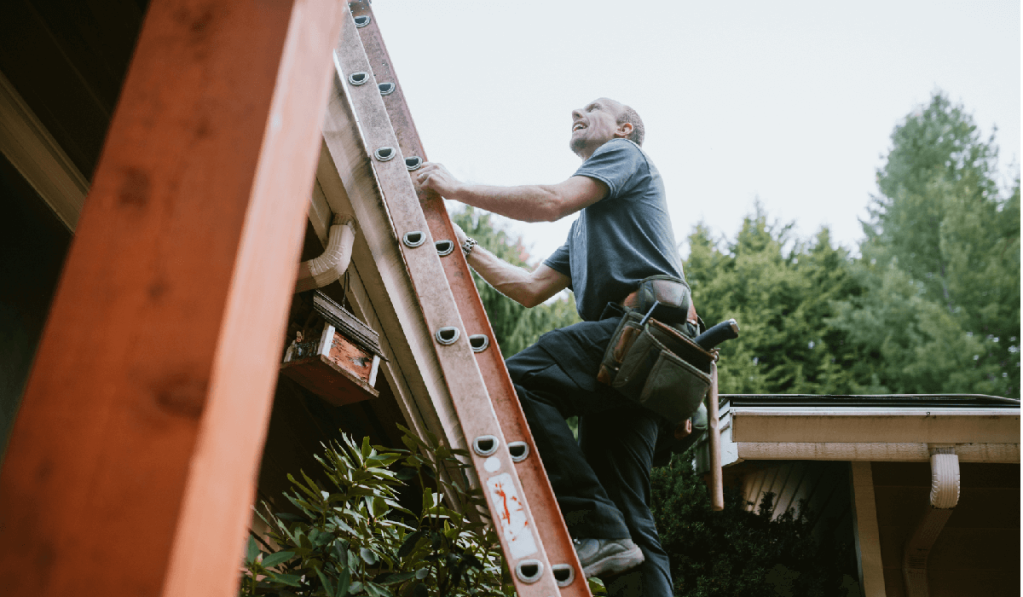 A man in a t-shirt climbs a ladder on the side of a house.