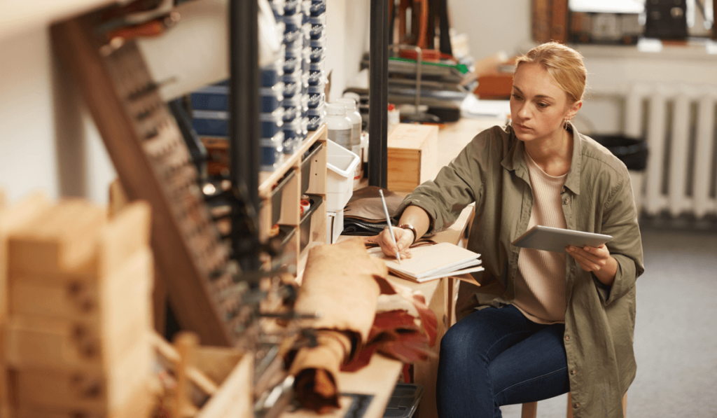 Woman at workbench holding one notebook while writing in another.