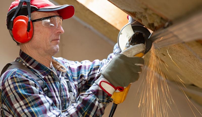 Man wearing blue plaid shirt with red safety headphones using a metal-cutting saw