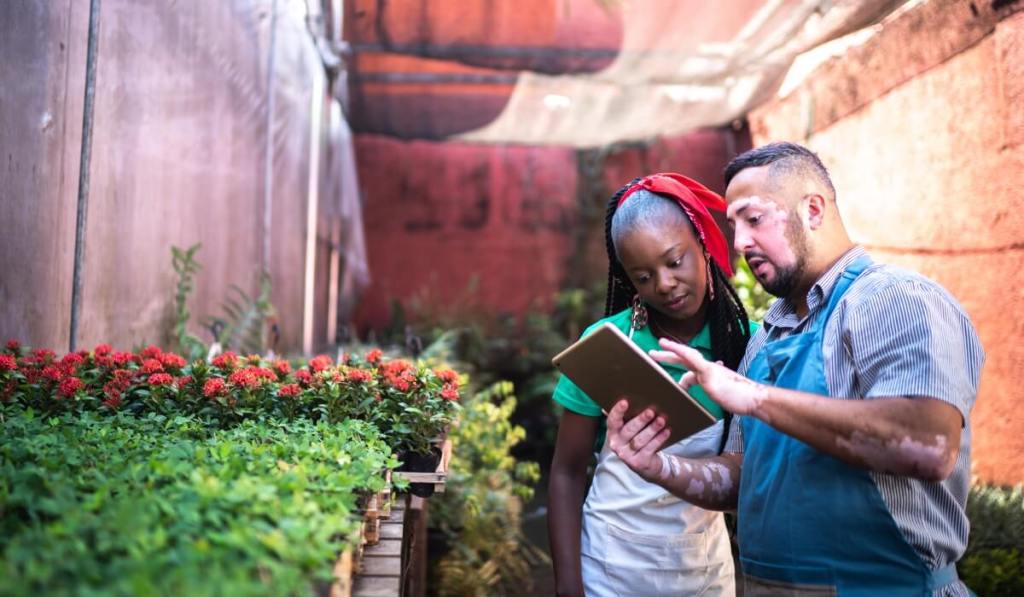 A man and woman stand in a greenhouse, looking over info on an iPad.