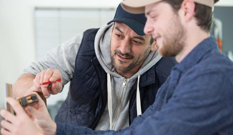 One man mentoring another man, both wearing baseball caps. One holds a measuring tape, the other holds a red pencil.