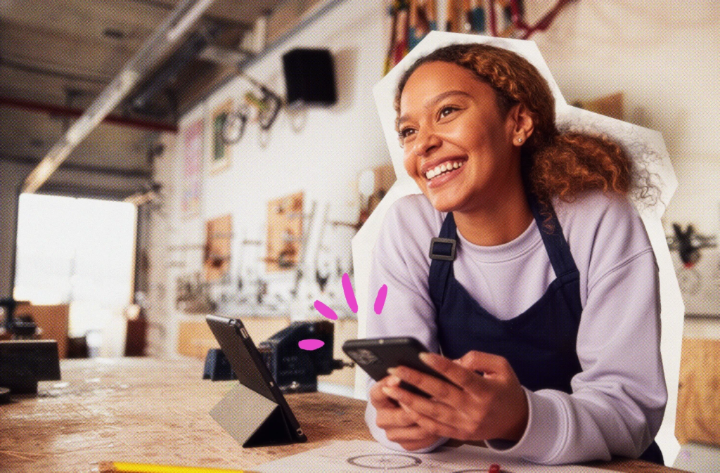 female presenting solopreneur with apronat work counter with card reader holding phone smiling