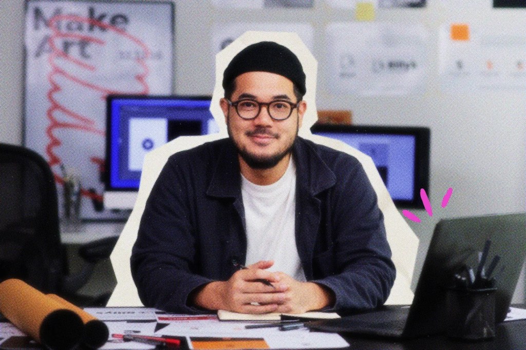 solopreneur sits at desk with laptop and work papers, holding a pen. they wear a beanie and glasses